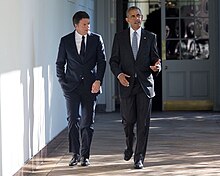 Obama and a man in a suit speak with walking down a hallway.