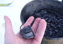 En hånd, der holder et stykke biochar med en spand af det i baggrunden