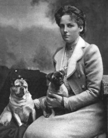 Black-and-white photograph of a serious-looking young woman in a suit, seated, with two small dogs