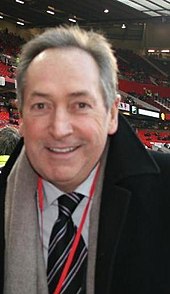 The upper body of a grey haired man. He is wearing a black coat, white shirt, black tie with blue stripes, grey scarf and a red ribbon is visible around his neck.