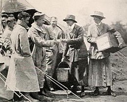 Eine Gruppe von Männern nähert sich zwei Frauen, alle in Uniform; Eine der Frauen hält eine große Kiste in der Hand. Sie sind im Freien und im Rahmen sind Zeltseile sichtbar.