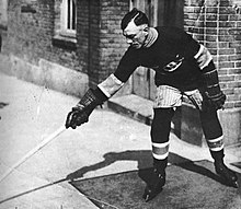 Svartvitt foto av Joseph Hall i hockeykugghjul utanför en byggnad.