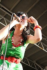 Katy Perry zpívající na koncertě, na sobě zelené šaty a růžové sluneční brýle s obručemi ve tvaru srdce