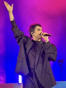 Grassi pictured on stage wearing black, with one arm outstretched and the other holding a microphone.