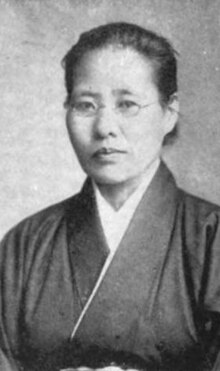 A black-and-white portrait of a middle-aged Asian woman, wearing glasses and a simple dark jacket. Her hair is dressed back, away from her face and neck.