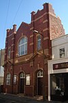 A tall building with deep red brickwork and yellowish stone dressings. There are two identical entrances: wooden doors in recessed stone doorways with prominent yellow keystones. A window to their left is in a similar recess. Two pediments flank a stone frieze with the words 