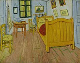 A small room with paintings on the wall, two chairs, a single bed and a table