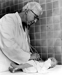 Dr. Virginia Apgar, standing, facing right, examining baby with stethoscope.