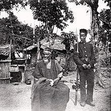Temne leader Bai Bureh seen here in 1898 after his surrender, sitting relaxed in his traditional dress with a handkerchief in his hands, while a Sierra Leonean West African Frontier Force soldier stands guard next to him Bai Bureh (1898).jpg
