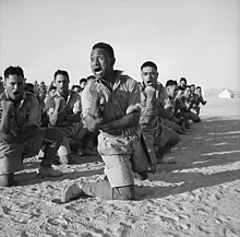 A squad of men kneel in the desert sand while performing a war dance