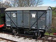 Ein aus Holzbohlen gebauter Kurzgüterwagen.  Die Seiten sind mittelgrau mit einem großen weißen G links von den Türen und einem großen weißen W rechts.