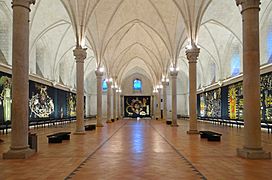 Photographie d'une salle voutée d'ogives où sont accrochées des tapisseries à dominante noir et or.