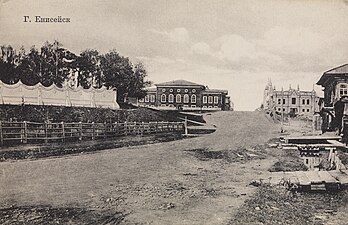 Photographie en noir et blanc d'un chemin de terre à la périphérie de la ville, avec des bâtiments en pierre et bois au fond de l'image.