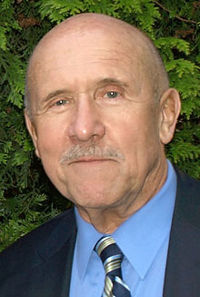 Middle-aged, balding white male with moustache, wearing suit and tie, smiling at camera