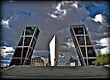 A stone monument stands in the center. Two skyscrapers leans to the center. The sky has been ominously altered.