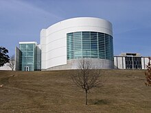 A large white and cement building sits on top of a large hill. A large round part of the building juts out from the rest and contains many windows.