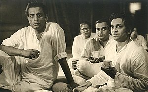 Photograph of Satyajit Ray seated with Ravi Shankar with several others in the background