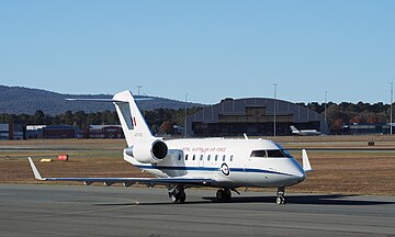 A twin-tailjet, high-tailplane passenger aircraft painted white above and grey below in front of an aircraft hangar and other buildings. A similar aircraft is visible in the background.