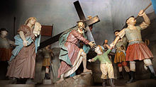 Photographie en couleurs. Groupe de statues colorées représentant Jésus montant une côte en portant une grande croix en bois, entouré de la foule.