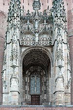 Portal de entrada de uma igreja de tijolos encimada por alto dossel de pedra e muito ornamentada com numerosas esculturas.