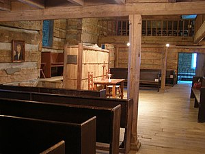 Fotografie des Inneren einer alten Blockkirche mit einer tragenden Säule aus grob behauenem Holz in der Nähe der Bildmitte. Die Säule trägt einen Holzbalken. Andere Balken sind sichtbar, die einen Balkon tragen, der den Raum an drei Seiten umgibt. Das Foto zeigt auf einen Kommuniontisch an der Vorderseite der Kirche und wurde von der linken Seite des Raumes unter dem Balkon aufgenommen. Links und auf der anderen Seite des Raumes sind ebene Holzbänke zu sehen. Der Boden ist aus Holz. Links an der Vorderwand des Raumes ist ein Porträt von Thomas Campbell zu sehen.