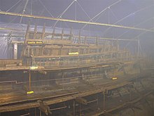 The deteriorated half of a wooden ship's hull. The view is from the port (left) side and is on the aft (back) half of the structure. The hull is in a large enclosed with sprinkler systems above it and scaffolding covered in plastic behind it. Each deck and some structural details ("sterncastle", "stern") are marked with large black signs with yellow text.