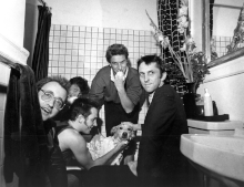 Fünf Männer in einem Badezimmer, das einen kleinen Hund umgibt, auf dessen Fell Shampoo verteilt ist.