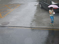 Uma mulher segurando um guarda-chuva é vista na calçada de uma rua durante uma chuva.