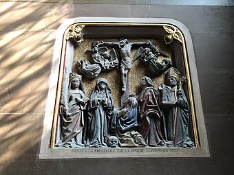 Groupe sculpté dans une niche représentant un homme sur une croix et, à ses pieds, trois femmes et deux hommes. L'une des femmes porte une couronne. L'un des hommes porte la mitre d'évêque et tient une maquette d'église. En dessous, lettres gravées en français.