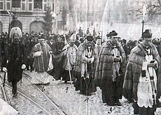 A imagem mostra um elevador de altos dignitários da igreja acompanhados por militares ou policiais.  Espectadores e companheiros de marcha podem ser vistos ao fundo.