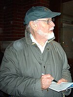 A man who is wearing a green cap and a dark green jacket is seen signing an autograph while look toward his left.