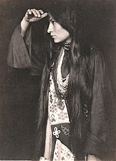 Woman with long hair standing in profile with hand to forward indicating she's looking far away