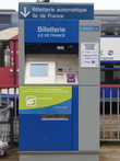 Appareil distributeur automatique de billets de couleurs bleue et grise portant l'indication « Billetterie automatique Île-de-France ». En arrière-plan, train de banlieue à deux niveaux.