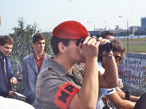 Red Beret-wearing, British, Royal Military Police member uses field glasses to look across the Berlin Wall from a viewing platform on the western side