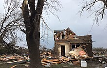 A home with severe damage with debris scattered around and large tree limbs snapped.