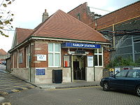 A brown-bricked building with a blue sign reading "FAIRLOP STATION" in white letters and a car parked in front all under a blue sky with white clouds