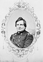 Black and white oval photo shows a round-faced, clean-shaven man. He wears a dark uniform with two rows of buttons and the eagle shoulder tab of a colonel.