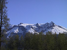 Uma montanha de topo plano e coberta de neve erguendo-se sobre árvores de folhas verdes em um dia claro.