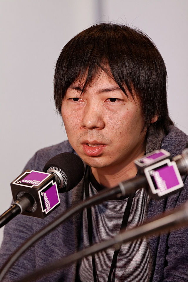 A photo of a middle aged Japanese man who is surrounded by microphones.