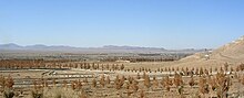 Spärliche braune Vegetation in staubiger Erde füllt den Vordergrund und verblasst zu fernen Bergen am Horizont.  Eine kaum wahrnehmbare Streuung von Gebäuden befindet sich im Mittelgrund.