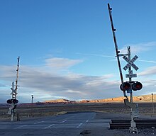 Railroad crossing signal on two-lane asphalt road with grassland in the midrange view and red cliffs and mesas in the distance