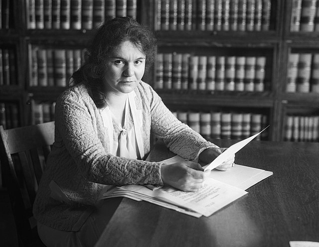 Une jeune femme à la peau claire et aux cheveux noirs, assise à une table, tenant des papiers, devant une bibliothèque remplie d'ouvrages juridiques.