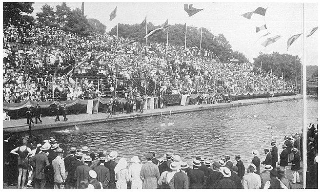 La piscine des Jeux olympiques : nageurs dans l'eau ainsi que sur les plages entourant le bassin, avec les juges-arbitres ; les tribunes remplies de spectateurs, au centre, la loge royale.