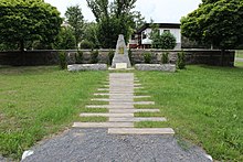 Ŝtona monumento, al kiu kondukas alirvojo el lignaj traboj. Malantaŭ la monumento muro kun arboj