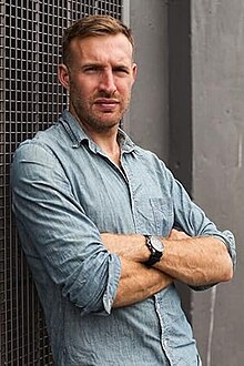 Photo of author Owen Laukkanen in a pale blue denim shirt leaning up against an industrial building in Vancouver, BC. His arms are crossed over his chest and his shirt sleeves are rolled up revealing a black watch. His brow is furrowed and he has a serious look on his face. His body is slightly angled away from the camera. His hair is light brown and short and he has short, neat facial hair.