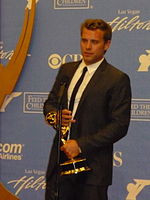 A man with dark hair, wearing a black suit, including a black tie and white T-shirt also holding a gold statuette