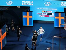 In der Totale sind zwei Kameraleute zu sehen. Michelle Coleman trägt beim Einmarsch in die Halle eine blaue Trainingshose, eine weiße Trainingsjacke und eine gelbe Badekappe.