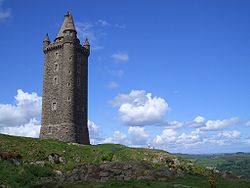 A photograph of a square tower in dark stone against a blue sky with white clouds