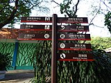 Un letrero etiquetado con direcciones que apuntan a un centro turístico, una clínica, la salida de un parque, una granja de animales para bebés, un jardín de pavos reales, un museo de zoología, un mundo de diversiones y un jardín de humedales.