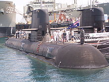 Un sous-marin à côté d'un quai, avec du personnel de la marine et des civils debout sur la coque extérieure.  Des parties d'un autre sous-marin et de deux navires de guerre sont visibles à l'arrière-plan.